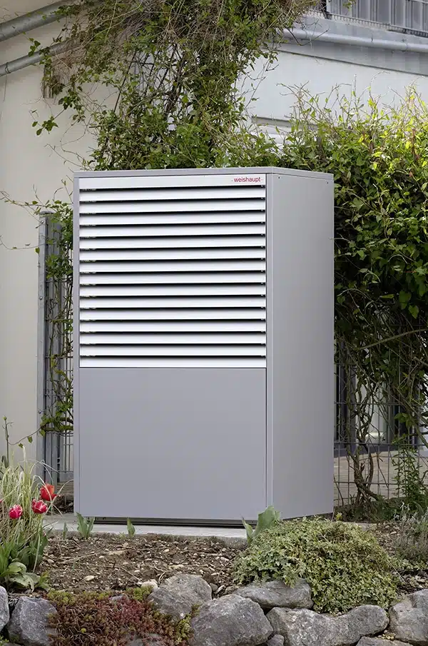 En grå propan varmepumpe med ventilationsgitre står i en have ved siden af en bygning.